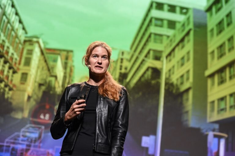 A woman wearing a ponytail and a black leather jacket stands on a stage. She looks slightly away from the camera. She wears a microphone and appears to be speaking to an audience.