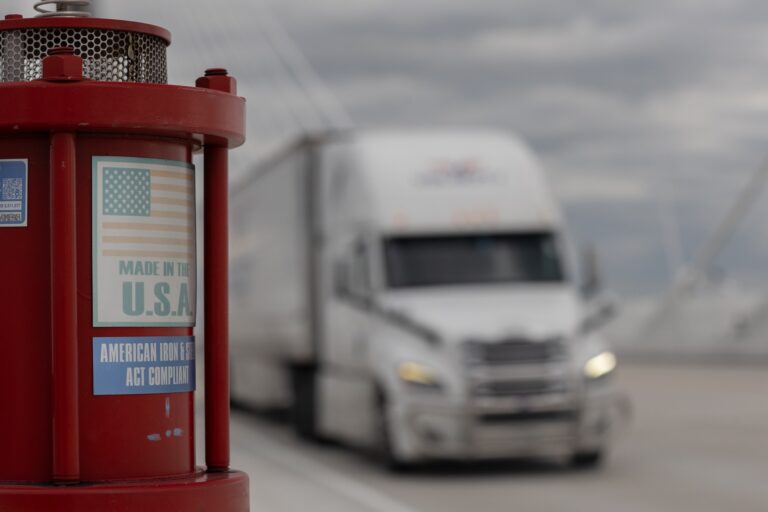 Close-up of a red American roadside call box marked “Made in the U.S.A.,” with a blurred semi-truck approaching on Gordie Howe International Bridge.