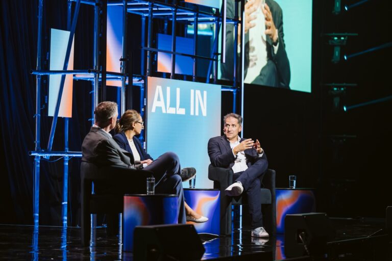 An image showing three people sitting on a stage. One of the people is wearing a suit jacket and a white button-down shirt, and appears to be speaking to the other two. Behind them, the words "All In" are visible on a blue screen in white letters.