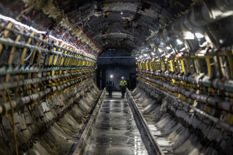 A shot looking down the length of a tunnel with conduits along its sides and railroad-style tracks on its floor. There are two workers in the distance wearing hardhats and headlights and walking toward the camera.
