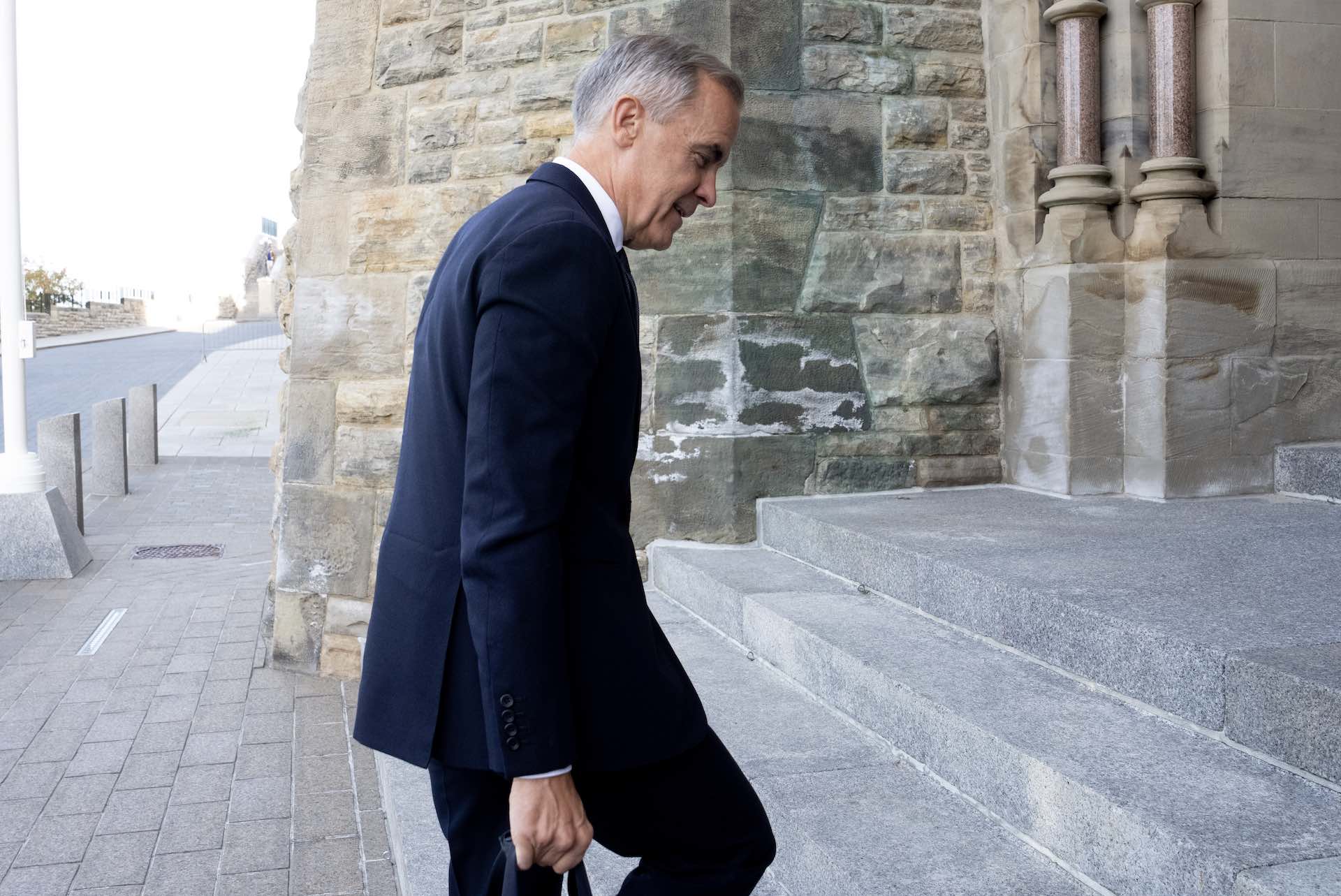 A side shot of Mark Carney walking up a set of stone steps into the Parliament buildings. He's wearing a dark suit. The straps of a case or tote bag are visible in his right hand.