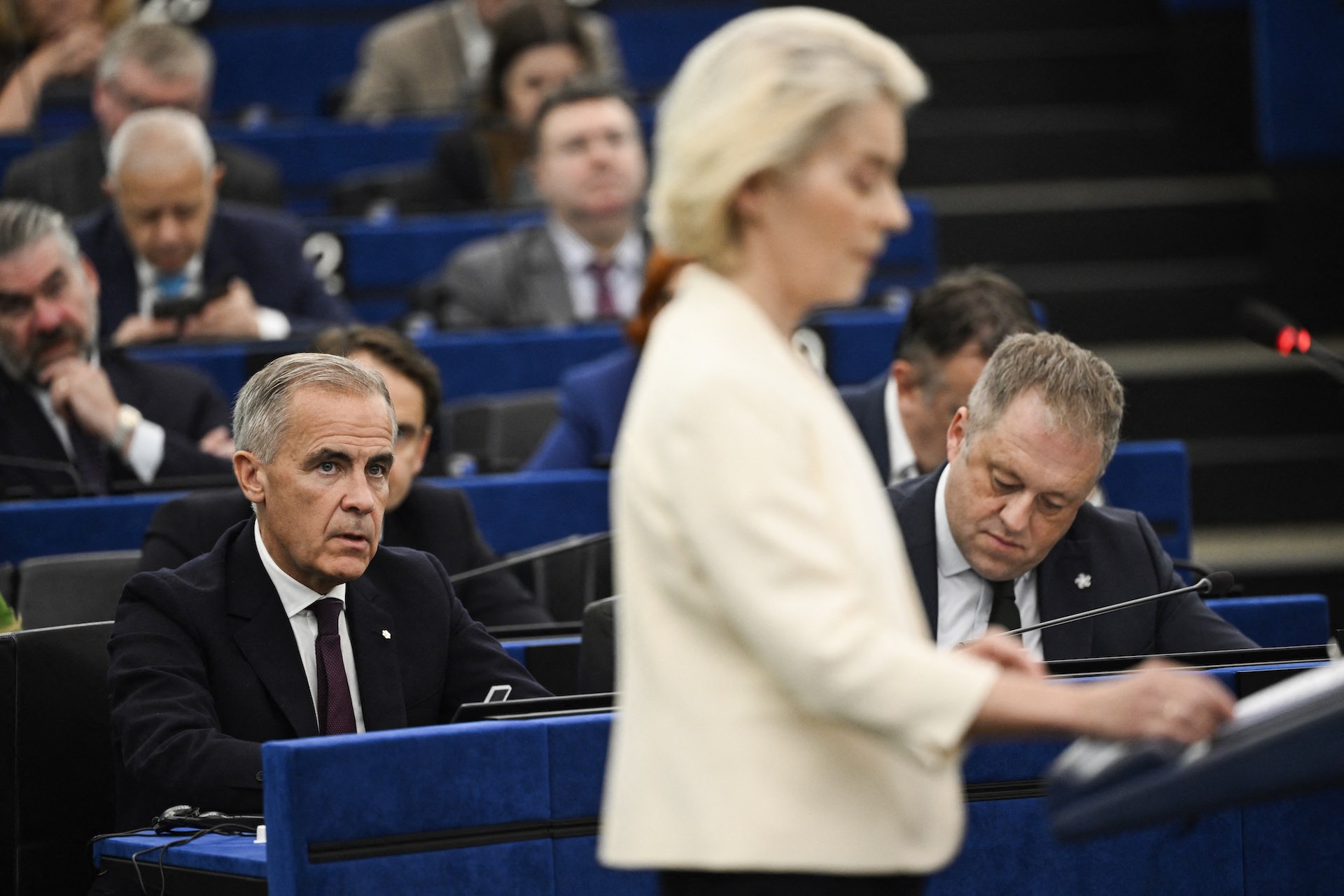 A shot of Mark Carney, who is seated, watching Ursula von der Leyen speaking at a lectern before the European Parliament. Carney is in sharp focus; von der Leyen is out of focus in the foreground.
