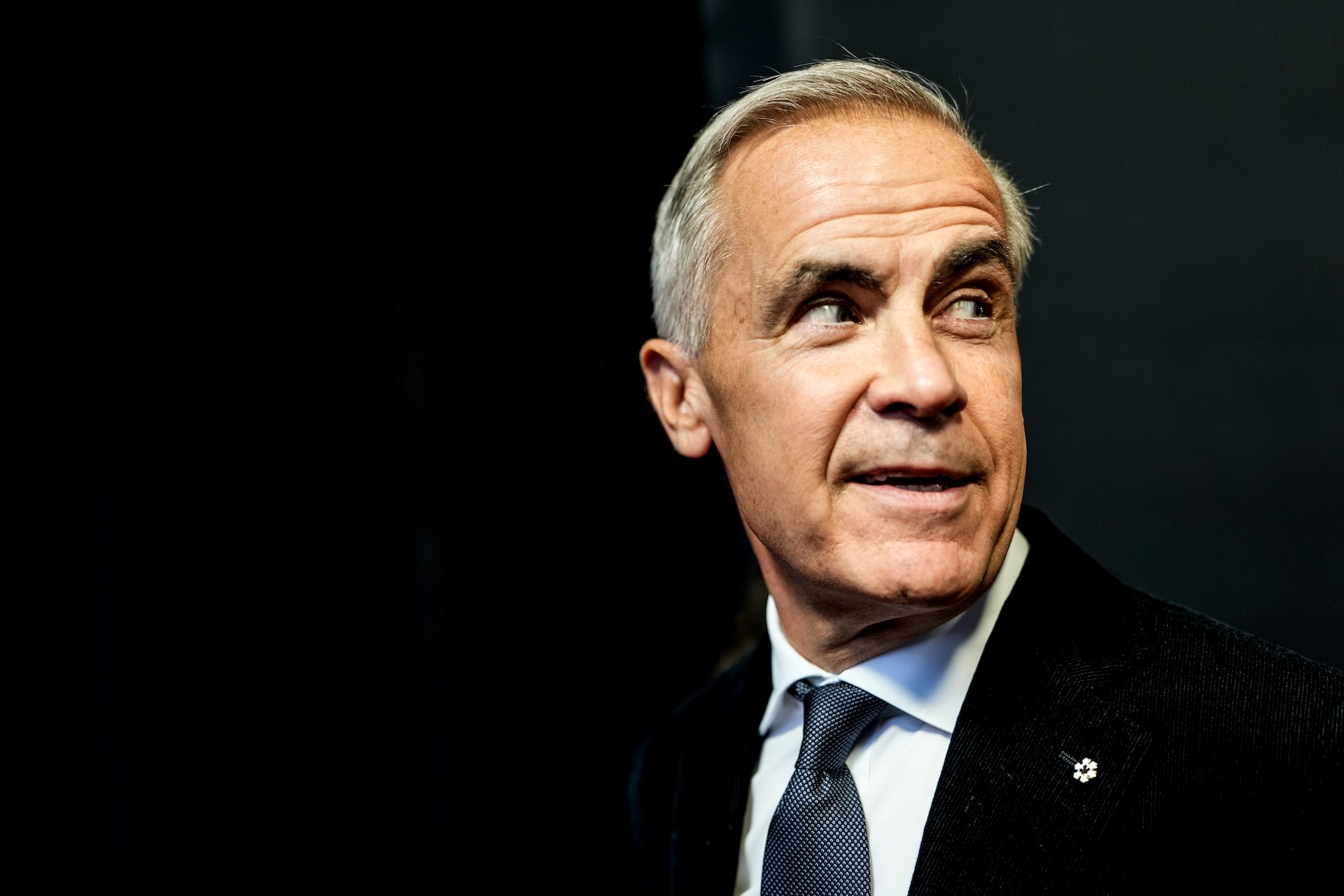 Close up of Mark Carney in a black suit against a black backdrop, looking over his shoulder with an eyebrow raised.