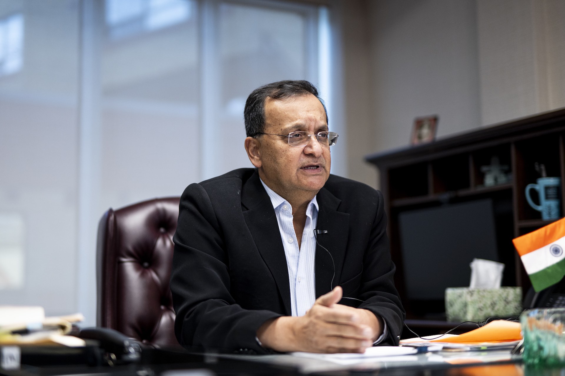 A shot of Dinesh Kumar Patnaik speaking to an interview while seated at an office desk. He's wearing a jacket and open-necked shirt and there is a small Indian flag among the objects on the desk.