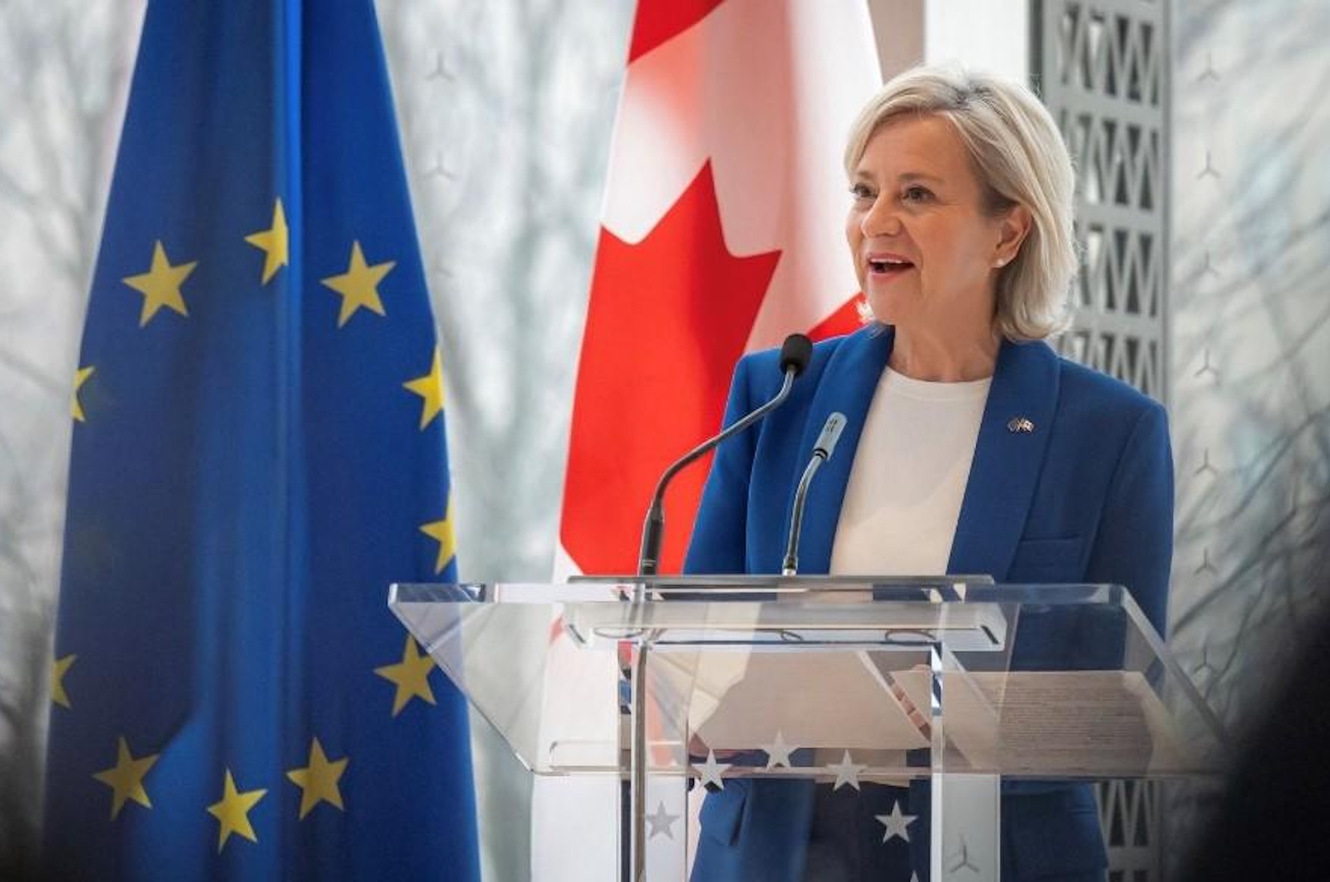 A shot of Genevieve Tuts speaking at a glass lectern, with the flags of the EU and Canada in the background. She's wearing a blue blazer.
