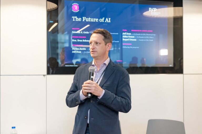 Jordan Jacobs stands holding a microphone. Behind him is a monitor showing the names of people on a panel under the heading "The Future of AI"