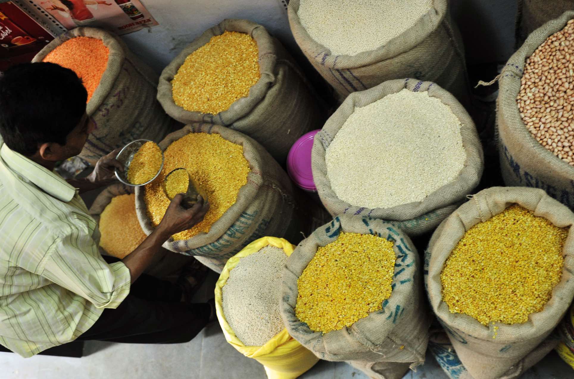 A shot of open sacks of lentils, beans and other dried foodstuffs in an outdoor market. There is a man on the far left side of the frame scooping lentils from one of the sacks into a dish.