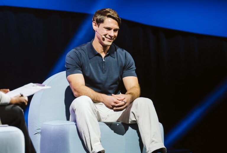 An image of a man wearing light pants and a blue collared shirt, sitting on a stage while wearing a microphone. He is smiling and looking at the ground.