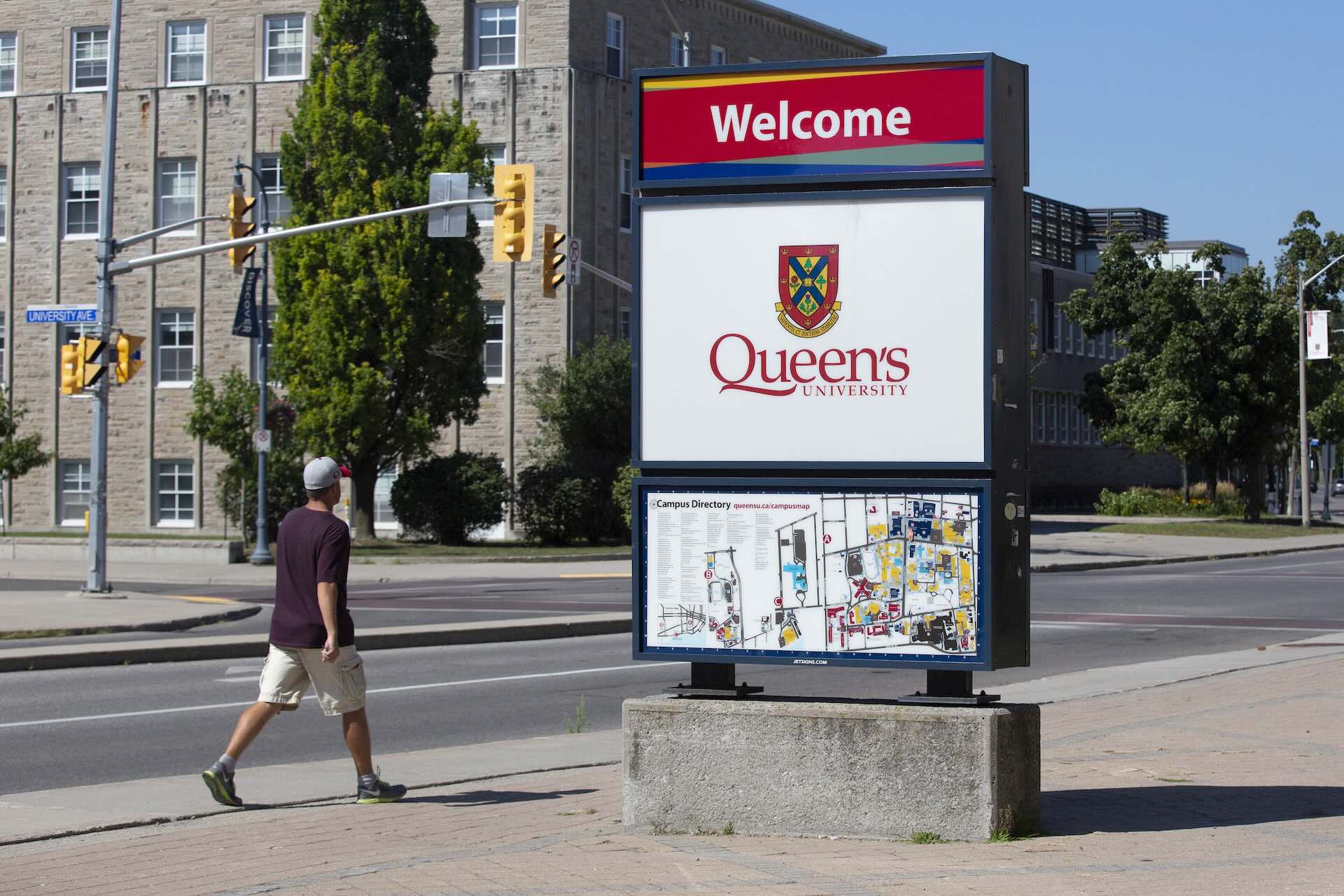 A Queen's University sign at the roadside near campus. There is a person walking by to one side of the sign.