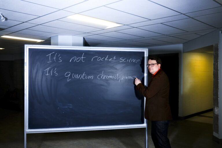 A man stands at a chalkboard while looking at the camera. He has apparently written "It's not rocket science; it's quantum chromodynamics."