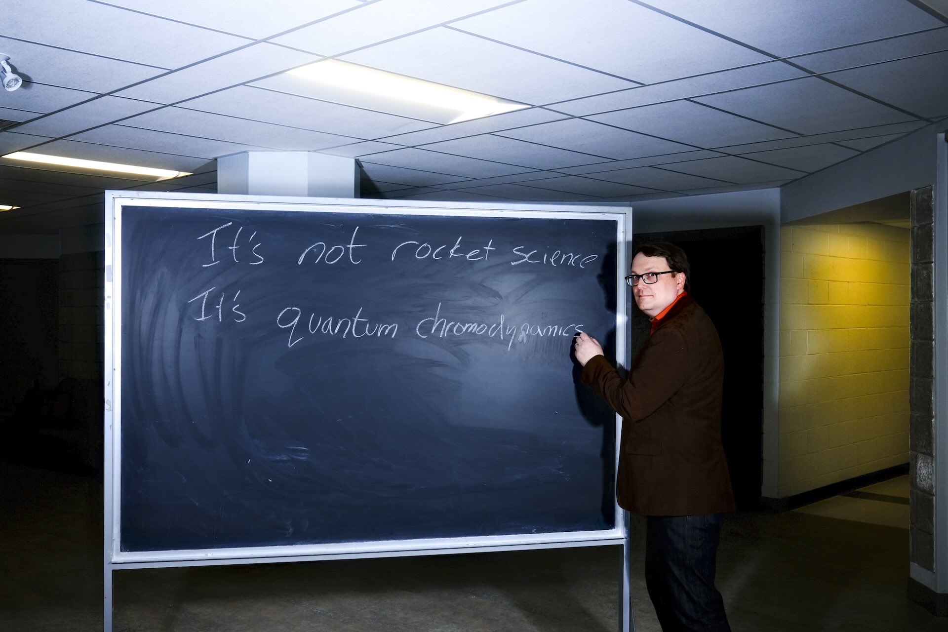 A man stands at a chalkboard while looking at the camera. He has apparently written "It's not rocket science; it's quantum chromodynamics."