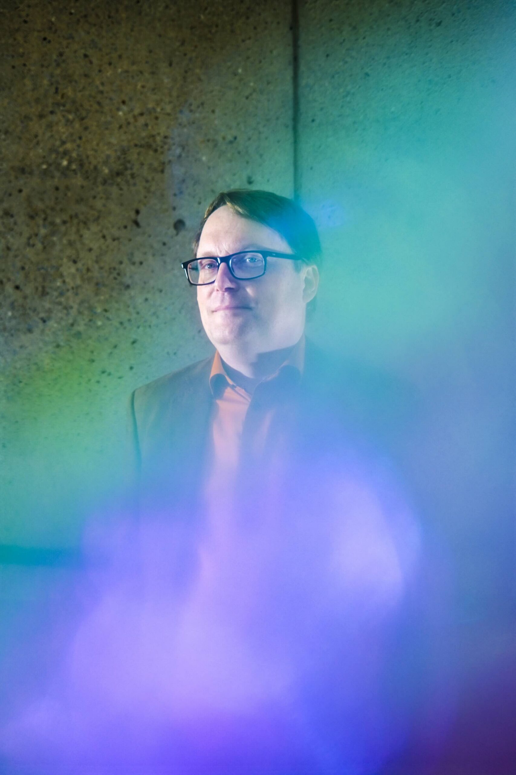 A man in glasses, a collared shirt and blazer looks at the camera, but is partially obscured by hazy blue and purple lighting
