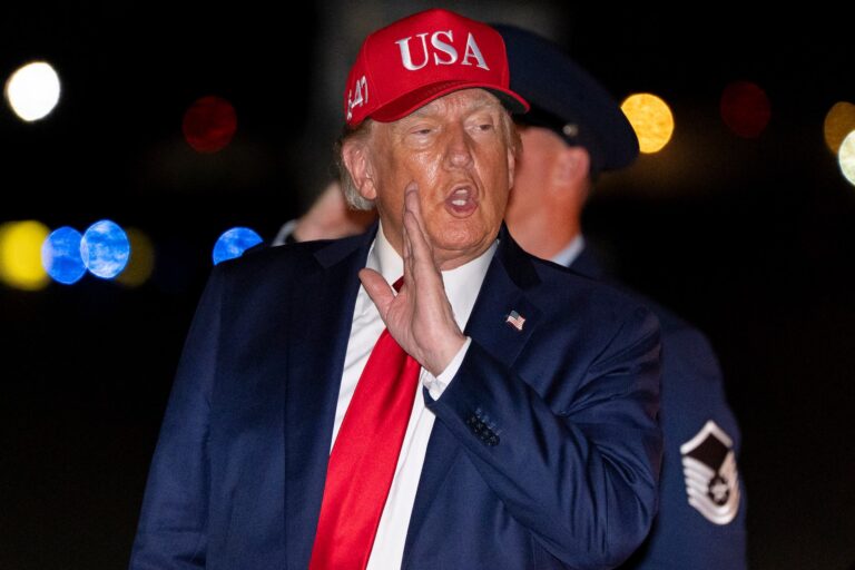 A nighttime shot of Donald Trump holding his hand while speaking. He's wearing a red hat with "USA" emblazoned on the front.