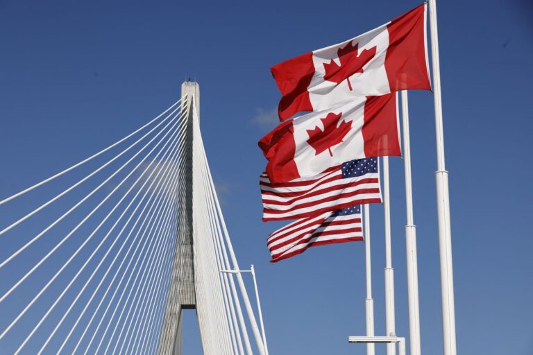 Canadian and U.S. flags wave beside Gordie Howe International Bridge under a clear blue sky.