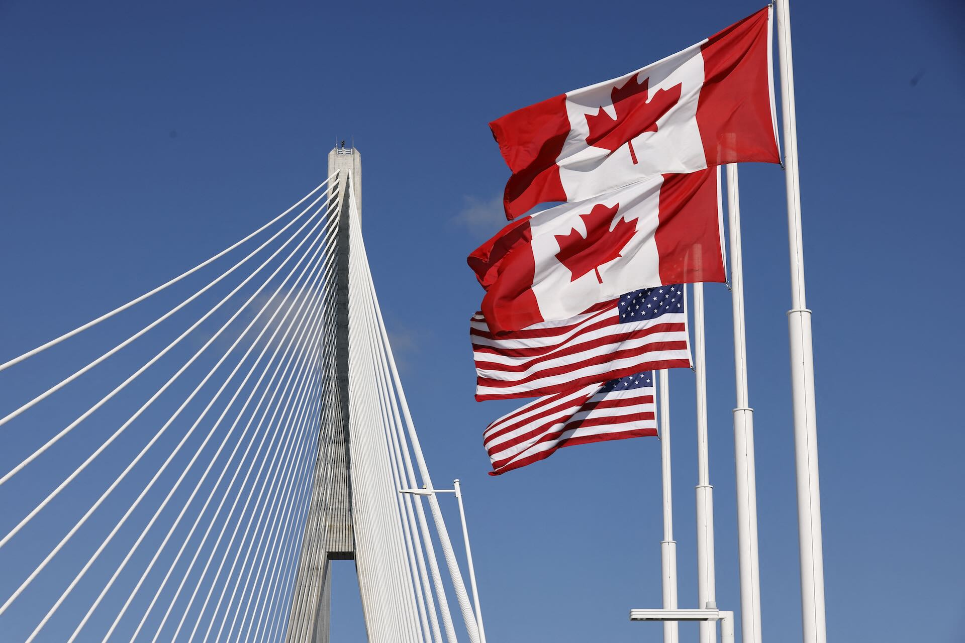 Canadian and U.S. flags wave beside Gordie Howe International Bridge under a clear blue sky.