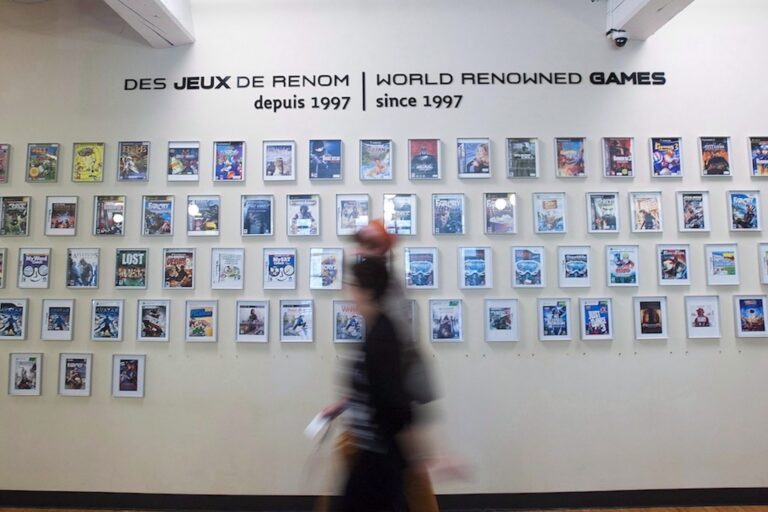 A shot of dozens framed video-game covers on a wall, with the label "World renowned games since 1997" inscribed above them. There are two passers-by blurred in the foreground.