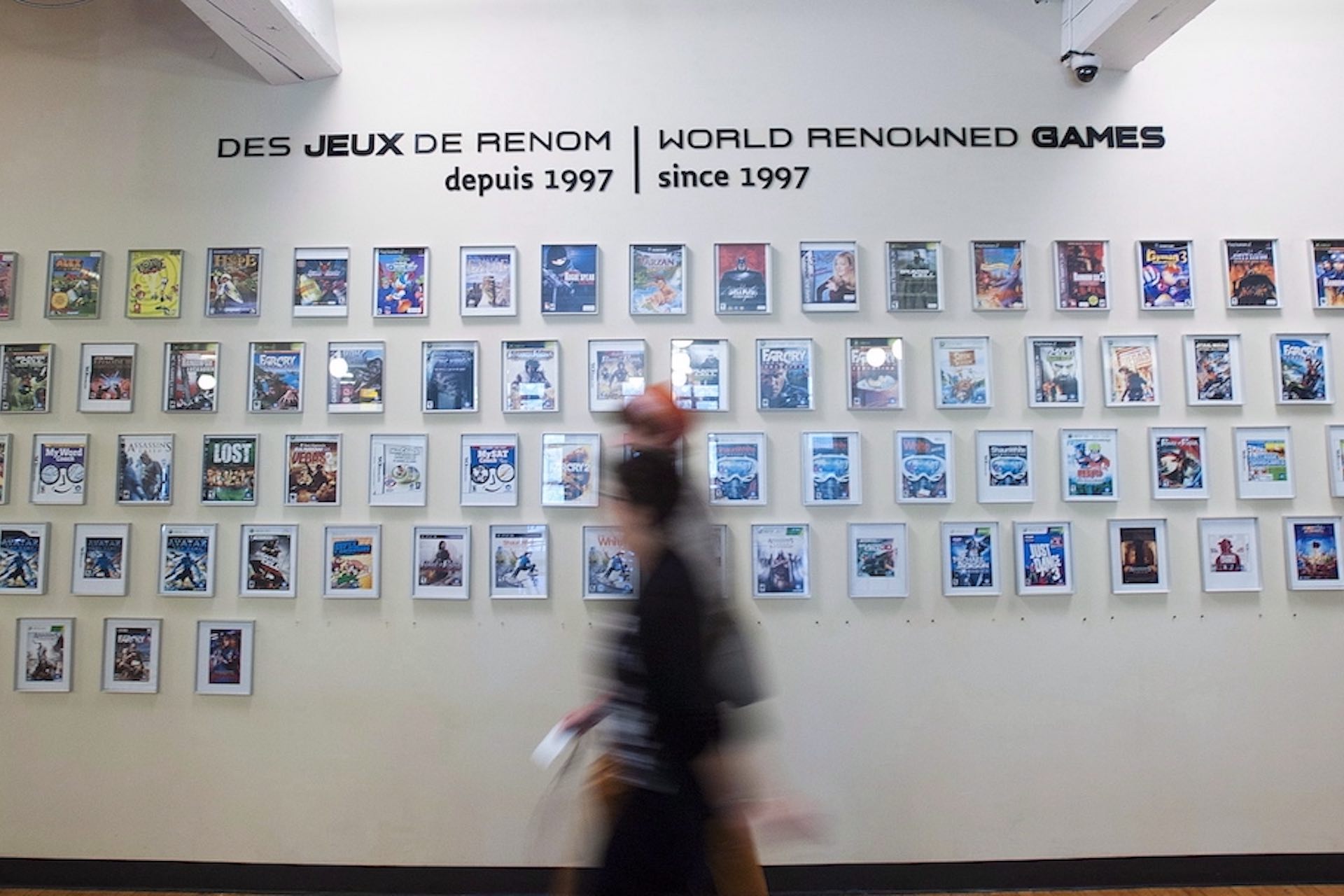 A shot of dozens framed video-game covers on a wall, with the label "World renowned games since 1997" inscribed above them. There are two passers-by blurred in the foreground.