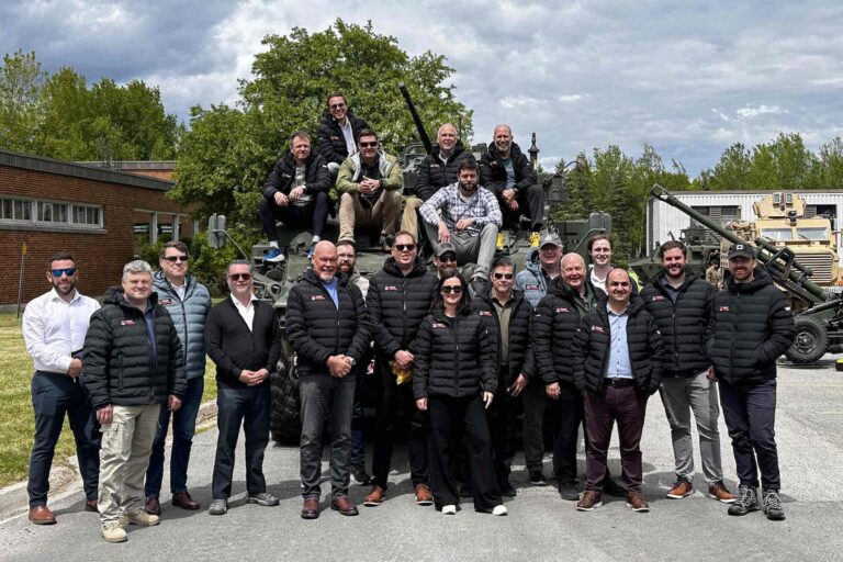 A group shot of people, many wearing branding black winter jackets, posed in front of an armoured vehicle. Several are sitting on the vehicle, in front of its gun turret.