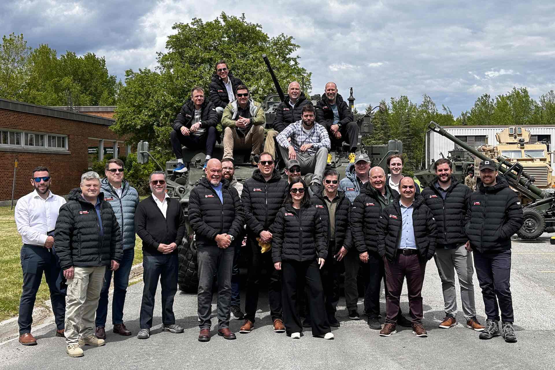 A group shot of people, many wearing branding black winter jackets, posed in front of an armoured vehicle. Several are sitting on the vehicle, in front of its gun turret.