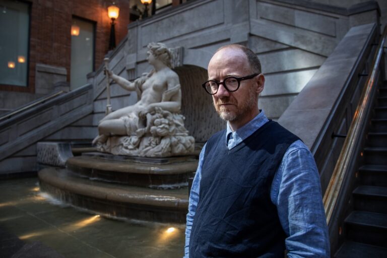 Willie Gagnon stands against a statue in a dimly lit building. He gazes off to the side,whiel wearing a black vest over a blue shirt.