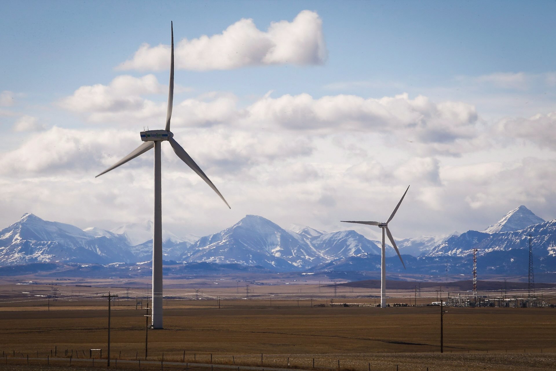 Wind turbines are shown standing tall in brown fields in the foreground. In the background are mountains and a blue sky with shite clouds.