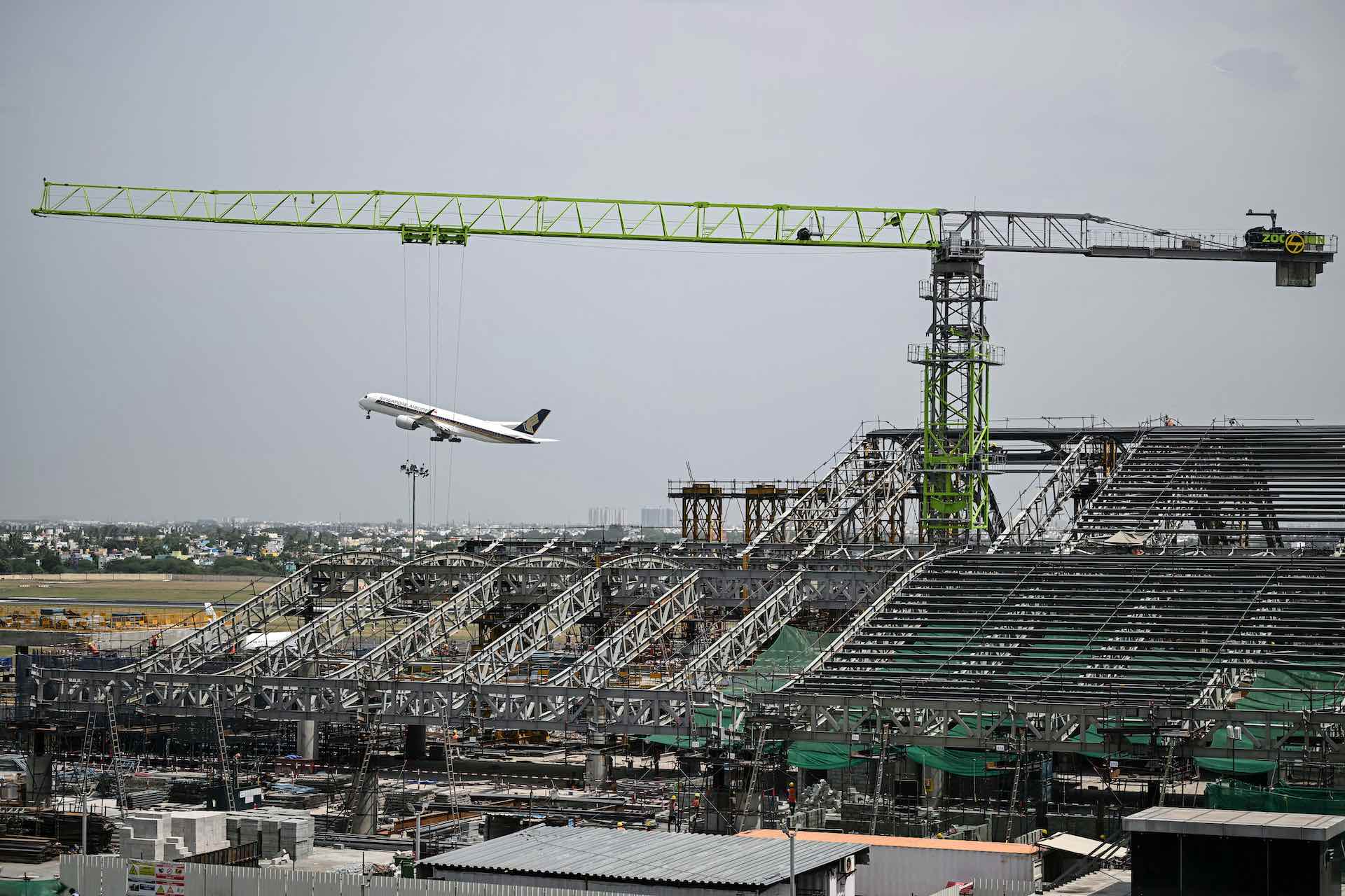 A wide shot of a construction site for a new airport terminal. There is a large green crane looming over the site, and a passenger jet taking off in the background.