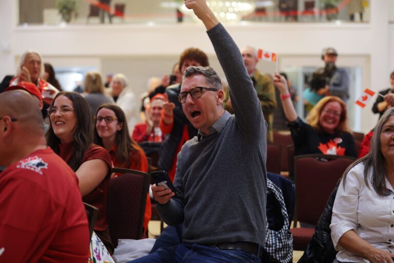 A photo of a man raising his arm and cheering among other pro-federalist supporters seated in rows of chairs in a large room. The man is wearing jeans and a grey sweater, but others in the frame are wearing red and waving small Canadian flags.