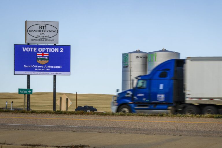 A photo of a blue roadside billboard taken in the rolling country of southern Alberta. The sign says, "Vote Option 2; Send Ottawa a Message." There is a transport truck speeding into the right side of the frame, with two grain silos beyond it.