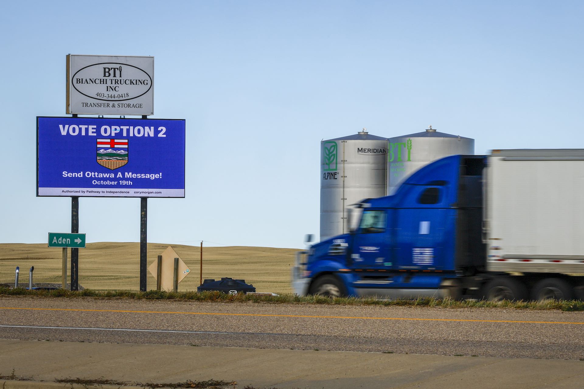 A photo of a blue roadside billboard taken in the rolling country of southern Alberta. The sign says, "Vote Option 2; Send Ottawa a Message." There is a transport truck speeding into the right side of the frame, with two grain silos beyond it.