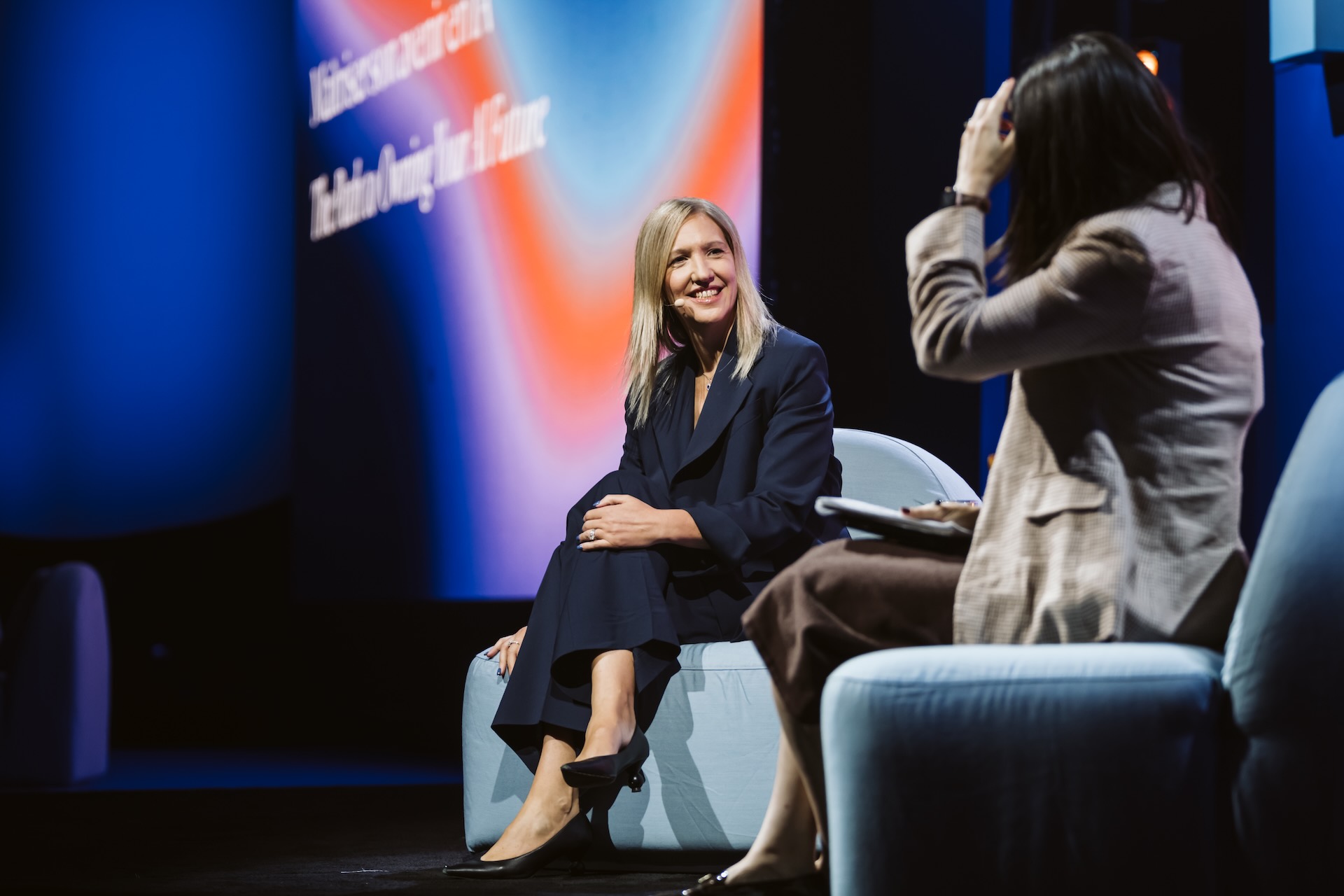 An image of two people sitting on a stage. A blonde woman sits facing the camera, smiling and wearing a dark pantsuit and heels. She is wearing a microphone.