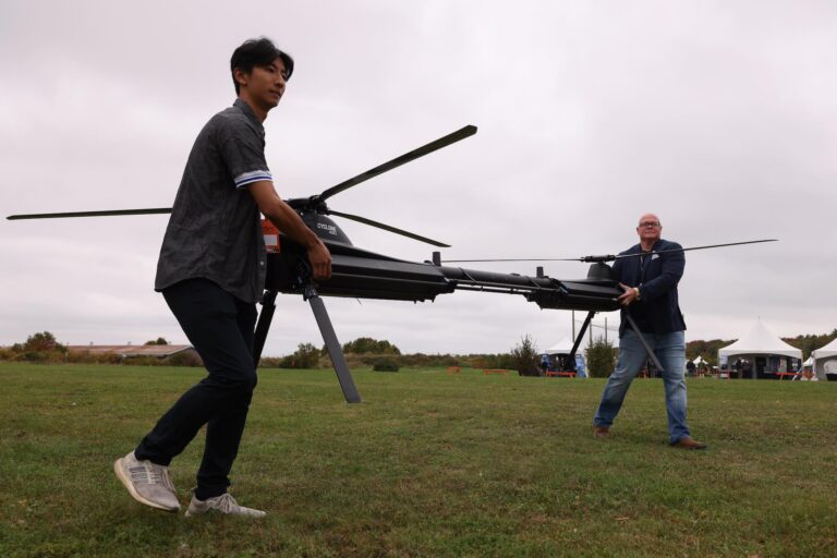 A wide frame of two men on field carrying a drone. The UAV looks to be about three metres long and is shaped like a bar with helicopter rotors on each end.