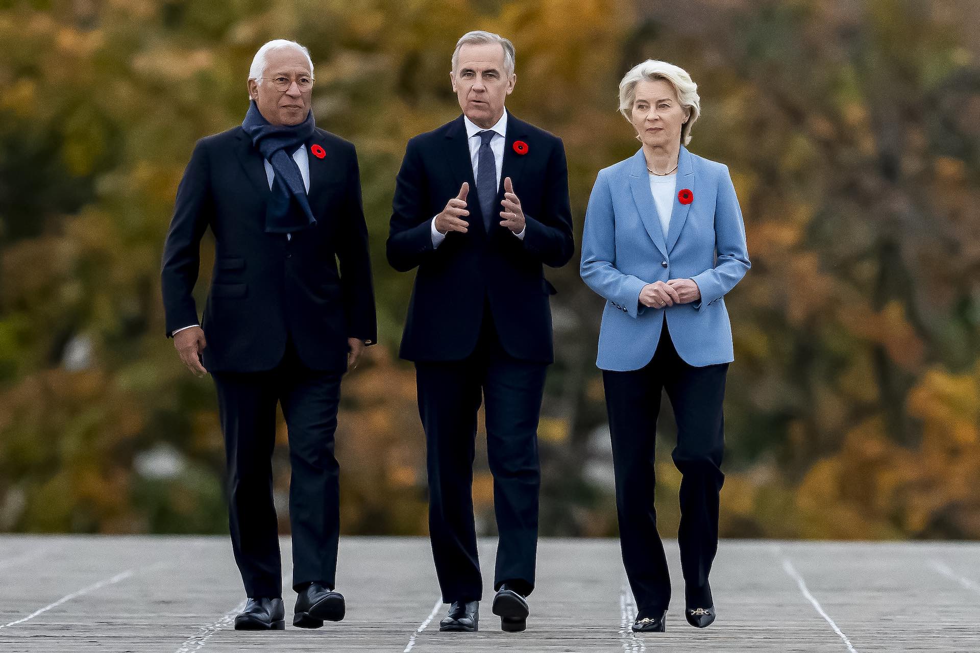 A head-on shot of, from left, Antonio Costa; Mark Carney; and Ursula von der Leyen walking across a deck. All three are wearing poppies, and autumn colours are visible on the trees in the background.
