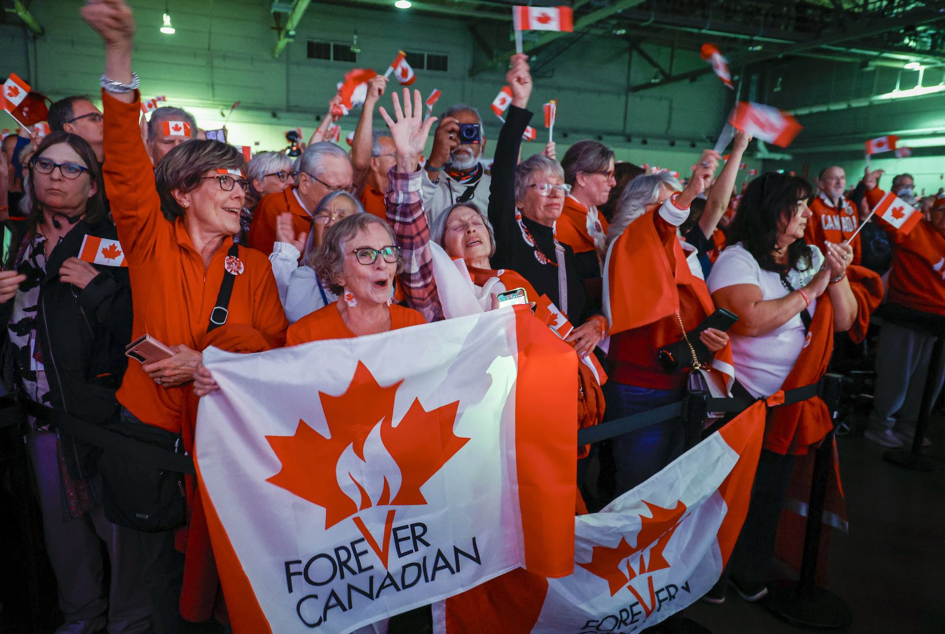 A shot of people at a rally cheering and waving paper Canadian flags. Those in the foreground are holding up flags that say "Forever Canadian." Many are wearing red.