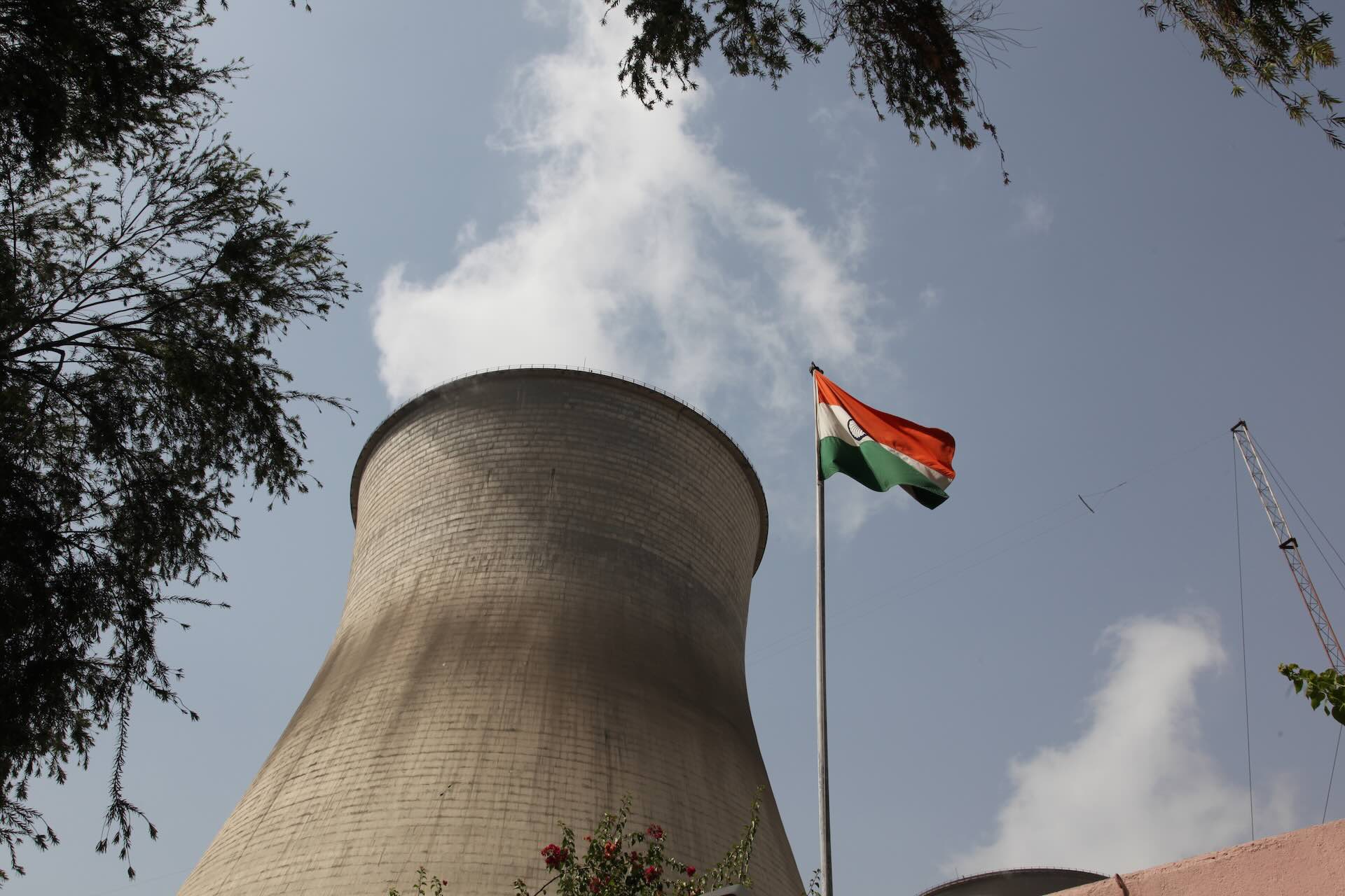 A low-angle photo of the cooling tower of a nuclear power station. There are tree fronds and an Indian flag in the foreground.