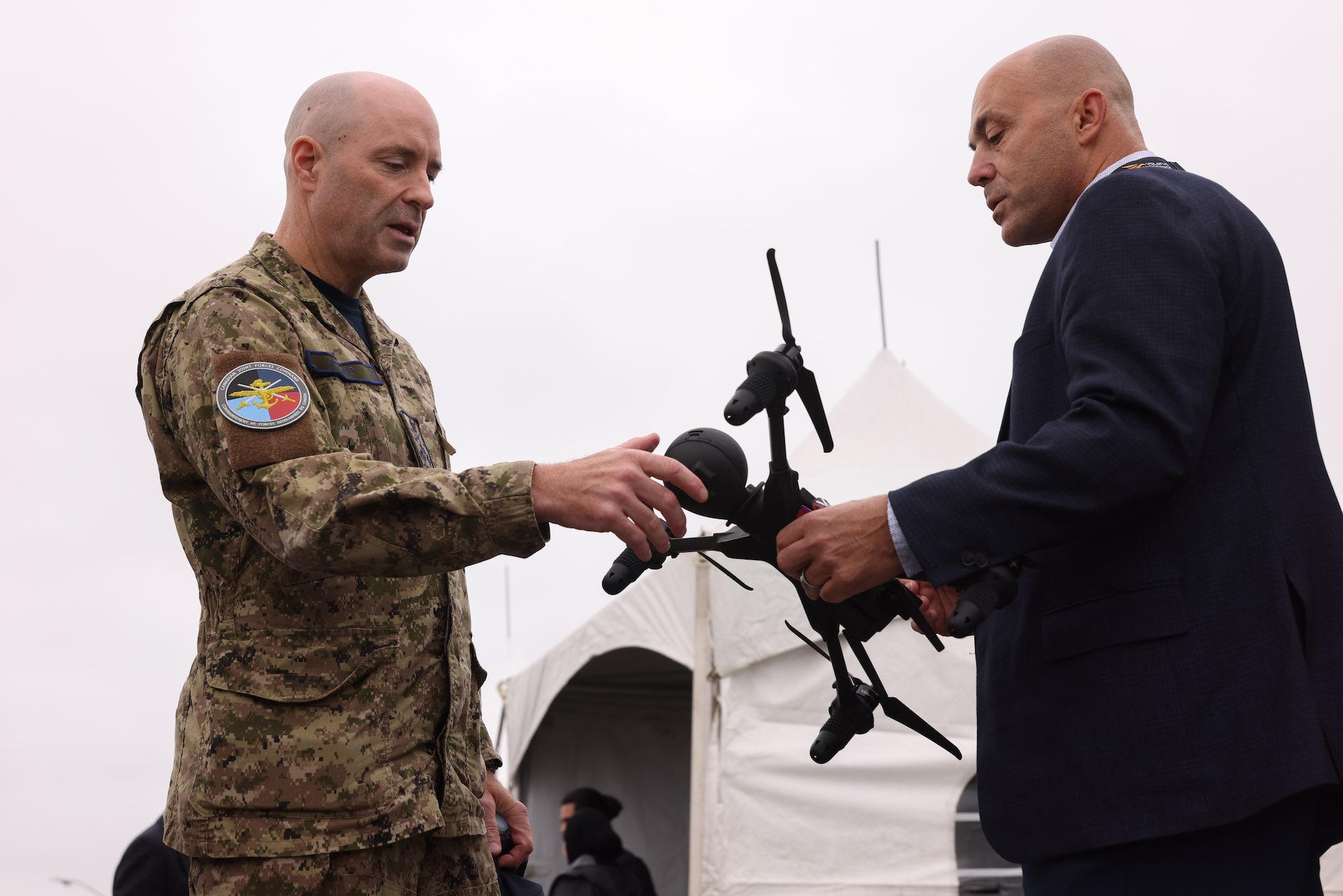 A photo of John Hryniw showing Lt.-Gen. Darcy Molstad a quadcopter drone. Hryniw is wearing a dark suit and Molstad is clad in in camouflage fatigues. There is a white tent in the background.