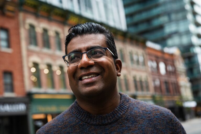 A man in glasses and a blue knit sweater outdoors on a city street, with brick buildings softly blurred behind him