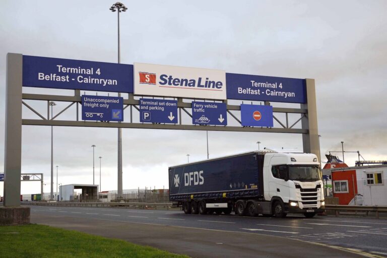 A shot of a freight truck passing beneath a gantry that bears signs directing traffic through the Port of Belfast.