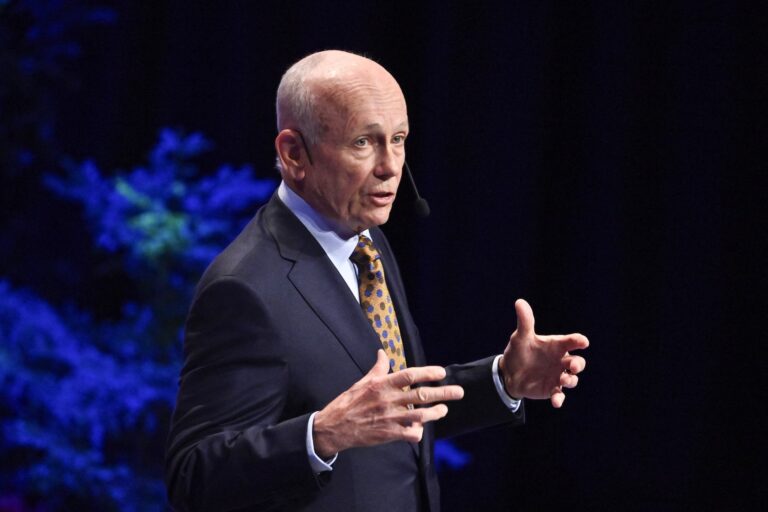A man wearing a dark suit jacket, blue button-down shirt and spotted brown-and-blue tie stands in front of a black and blue background, gesturing and speaking into a microphone.