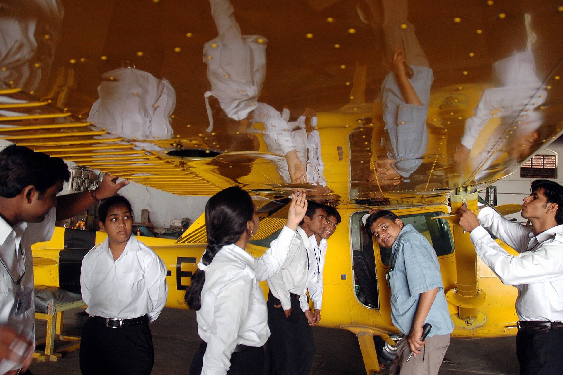 A shot of pilots-in-training, both men and women, under the wing of a small, yellow airplane. An instructor is opening the door of the aircraft.