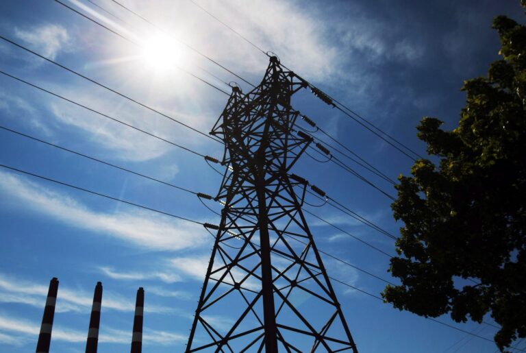 A low-angle shot of a pylon supporting high-tension power lines taken against the backdrop of sunshine and blue sky.