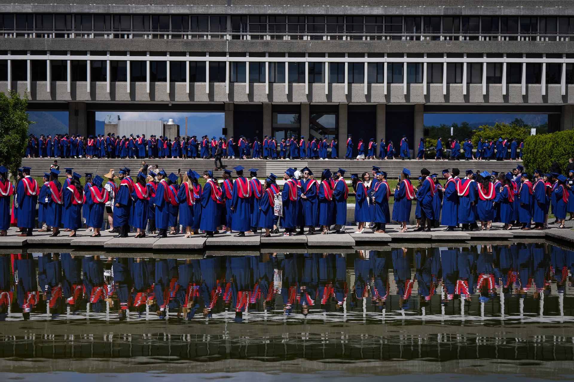 A shot of university grads lining up for convocation among the modernist concrete structures of Simon Fraser University. They're wearing blue caps and gowns with red hoods. There is a queue in the foreground beside a reflecting pool, and another in the background on a terrace.