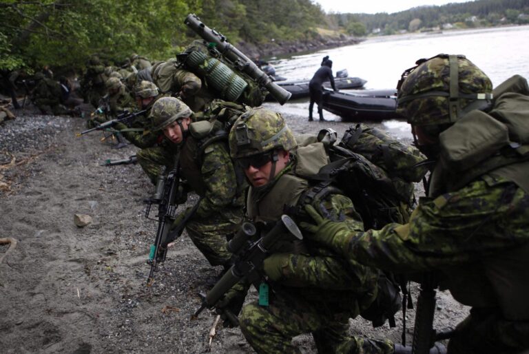 A side shot of a row of fully equipped soldiers making a beach landing from zodiac watercraft, rifles in hand.