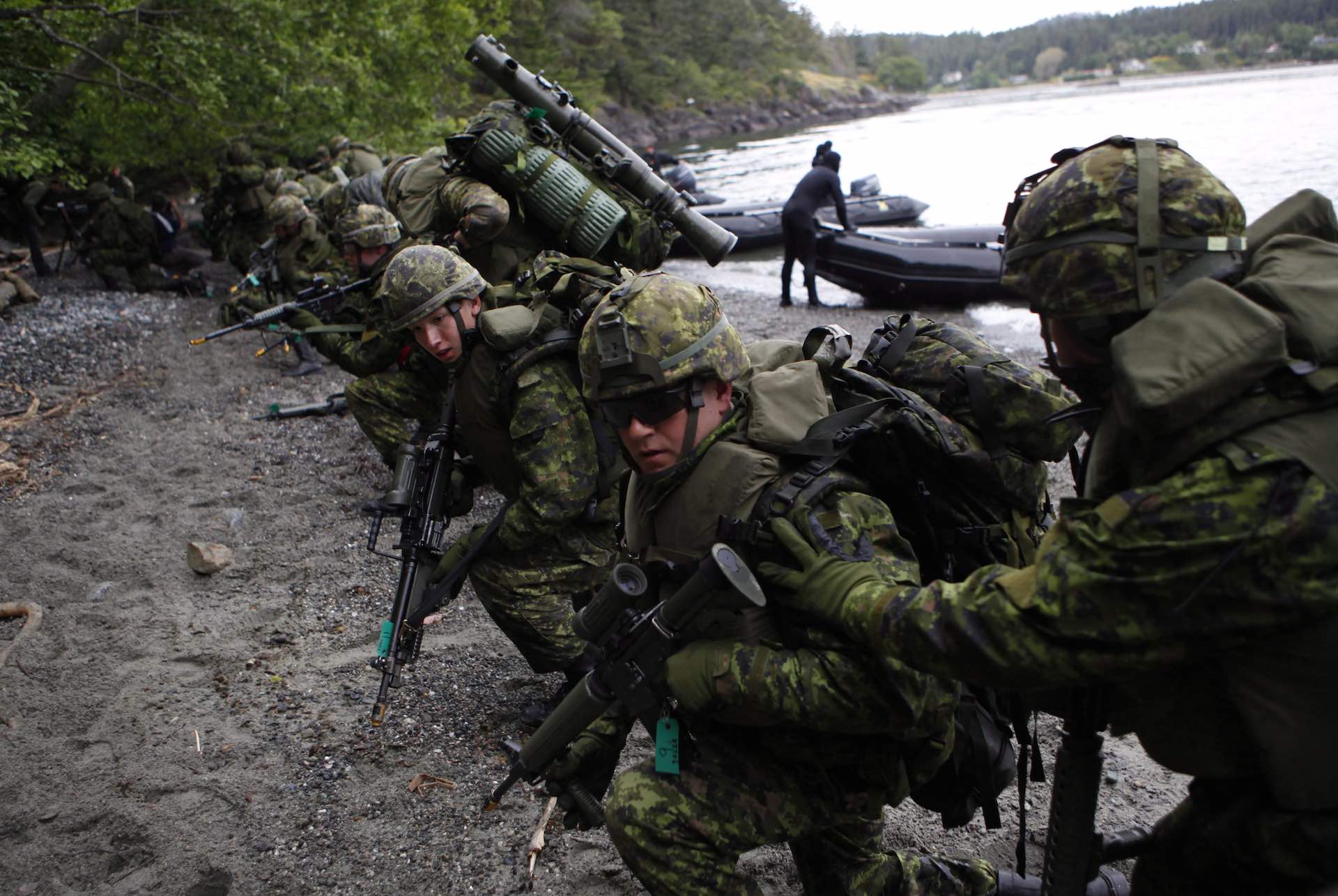A side shot of a row of fully equipped soldiers making a beach landing from zodiac watercraft, rifles in hand.