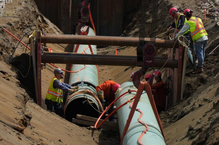 A shot of workers in a wide trench aligning light green-painted segments of pipe.