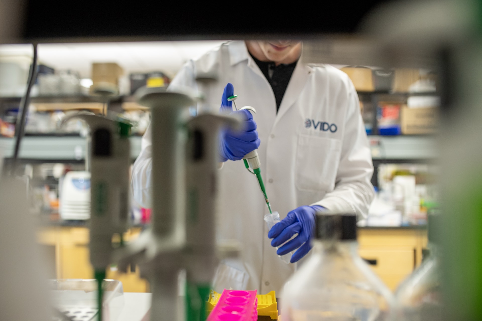 A shot taken through laboratory equipment of a researcher injecting liquid into a tube. The worker is wearing blue, latex gloves, and is a white lab coat with the VIDO logo on it.